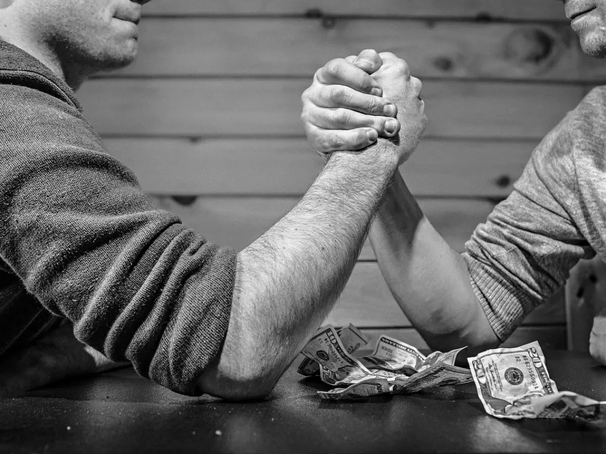 Two men arm wrestle over a pile of cash