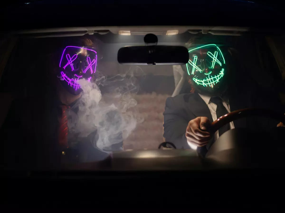 A car interior with two people wearing scary masks. The man in the driver's seat is looking directly at the camera.