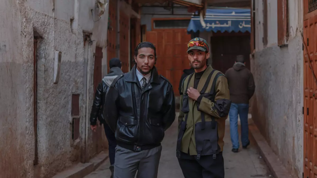 Two men stand in a narrow alleyway, one wearing a black leather jacket and the other in a green military jacket. The background is a gray wall with a wooden door.