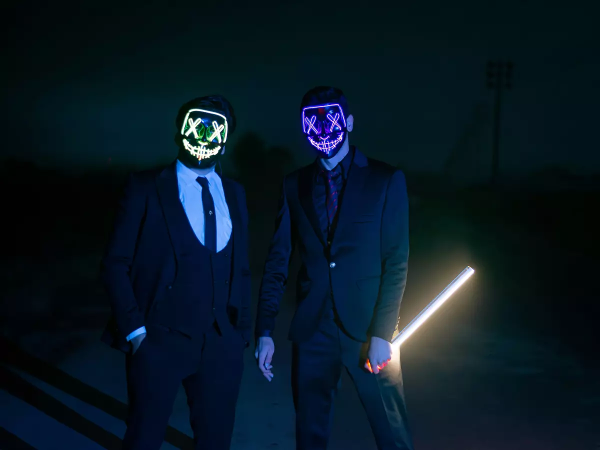 Two men wearing neon masks and suits standing in the darkness with one holding a white glow stick