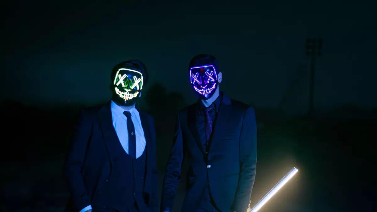 Two men wearing neon masks and suits standing in the darkness with one holding a white glow stick