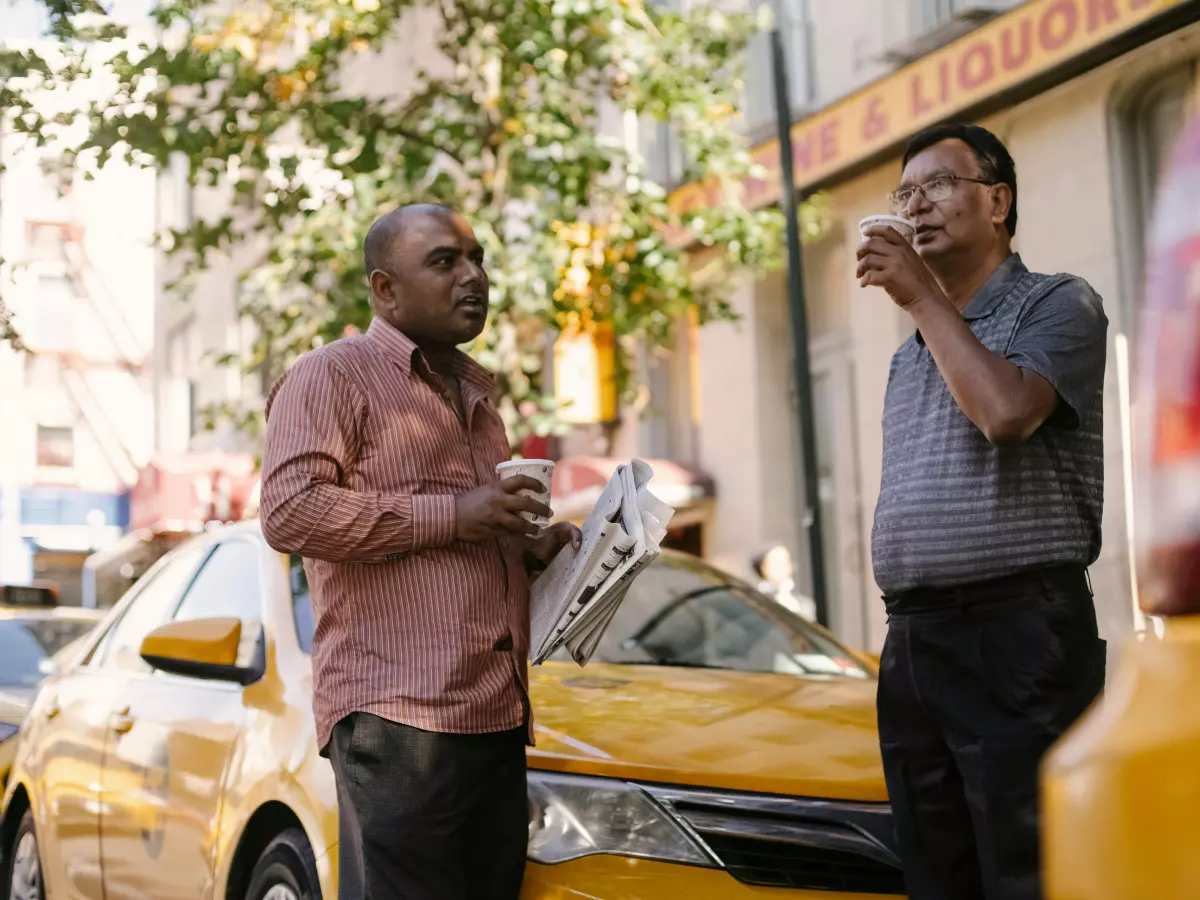 Two men are standing on a city street with a yellow taxi behind them. They are both looking at their phones, one is talking on his phone.