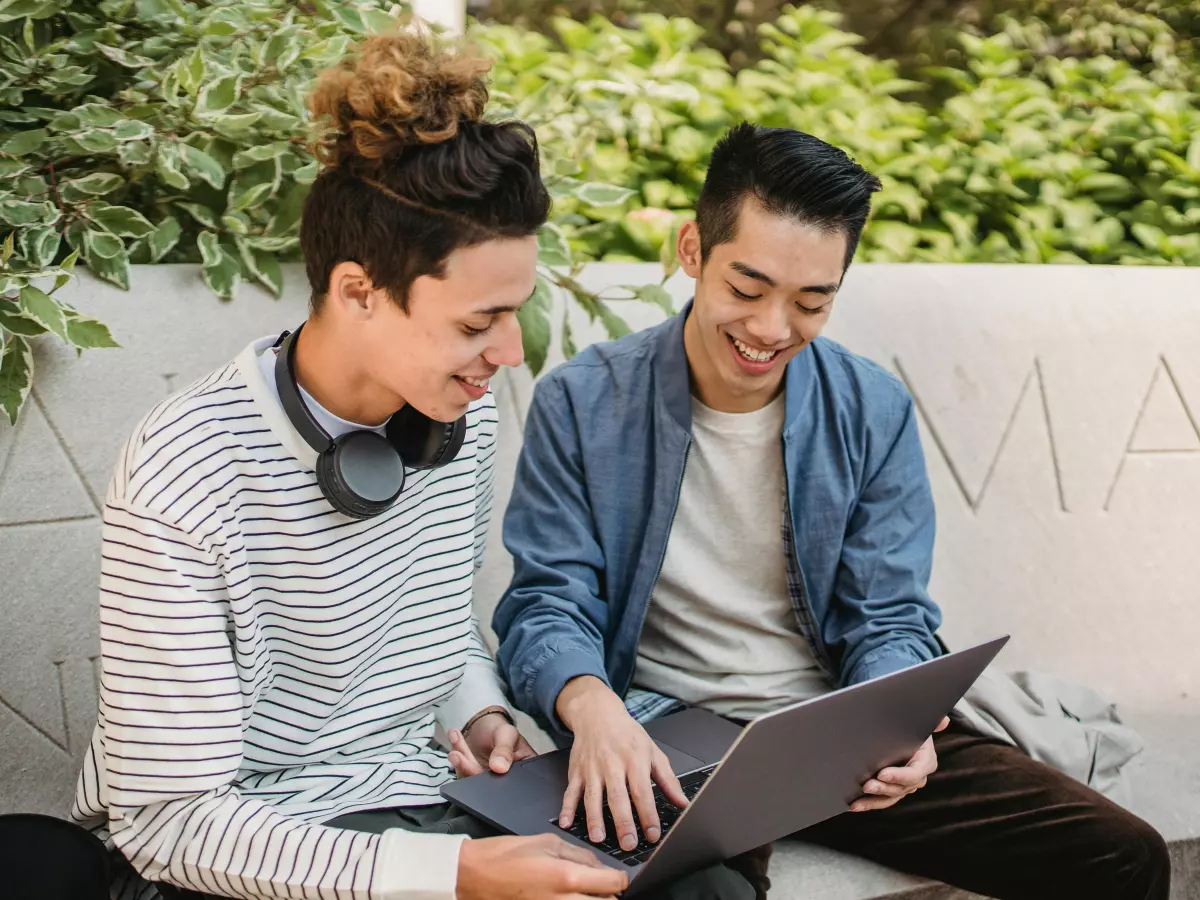 Two young men sitting on a bench outdoors, looking at a laptop screen. One man is wearing a striped shirt and headphones, the other is wearing a denim shirt.