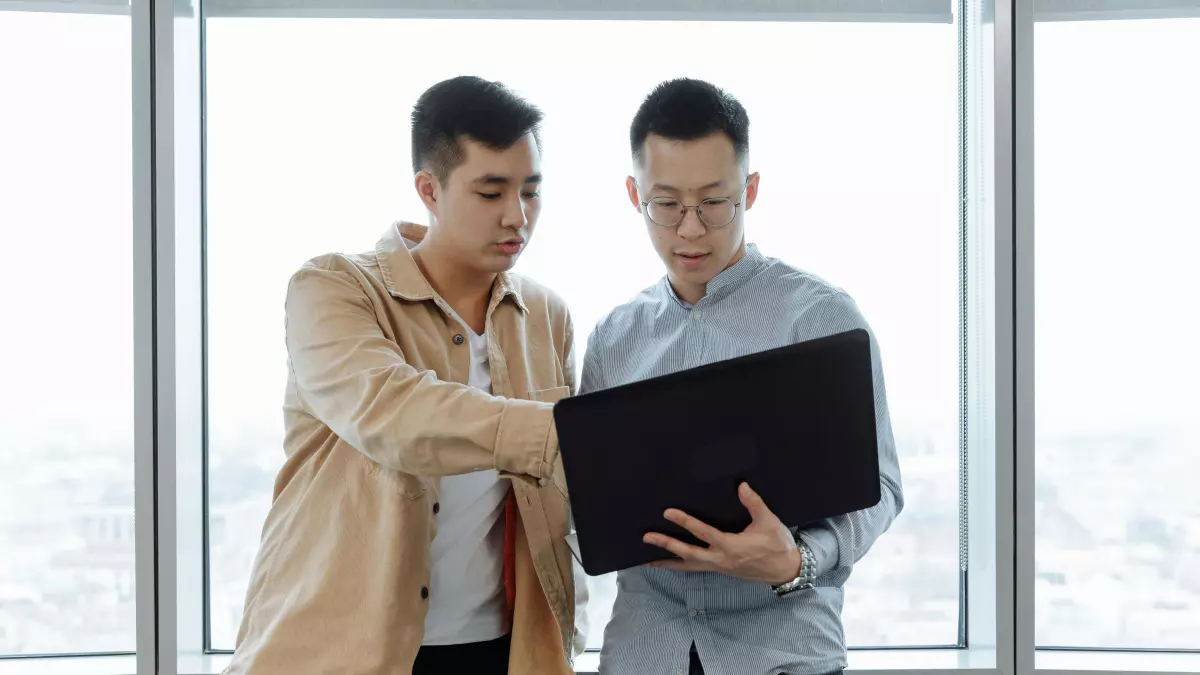 Two men, likely colleagues, collaborate at a desk, both looking at a laptop screen. The image conveys a sense of collaboration and teamwork in a professional setting.