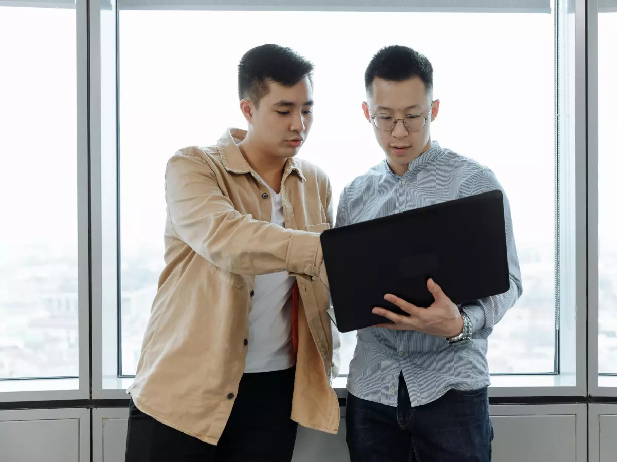 Two men, likely colleagues, collaborate at a desk, both looking at a laptop screen. The image conveys a sense of collaboration and teamwork in a professional setting.