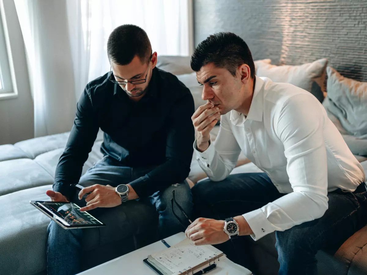 Two men, one in a black shirt and the other in a white shirt, are sitting on a bed. They are both looking at a tablet and a notebook. The man in the black shirt is holding a pen and is writing in the notebook.