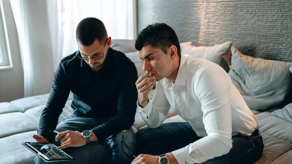 Two men, one in a black shirt and the other in a white shirt, are sitting on a bed. They are both looking at a tablet and a notebook. The man in the black shirt is holding a pen and is writing in the notebook.