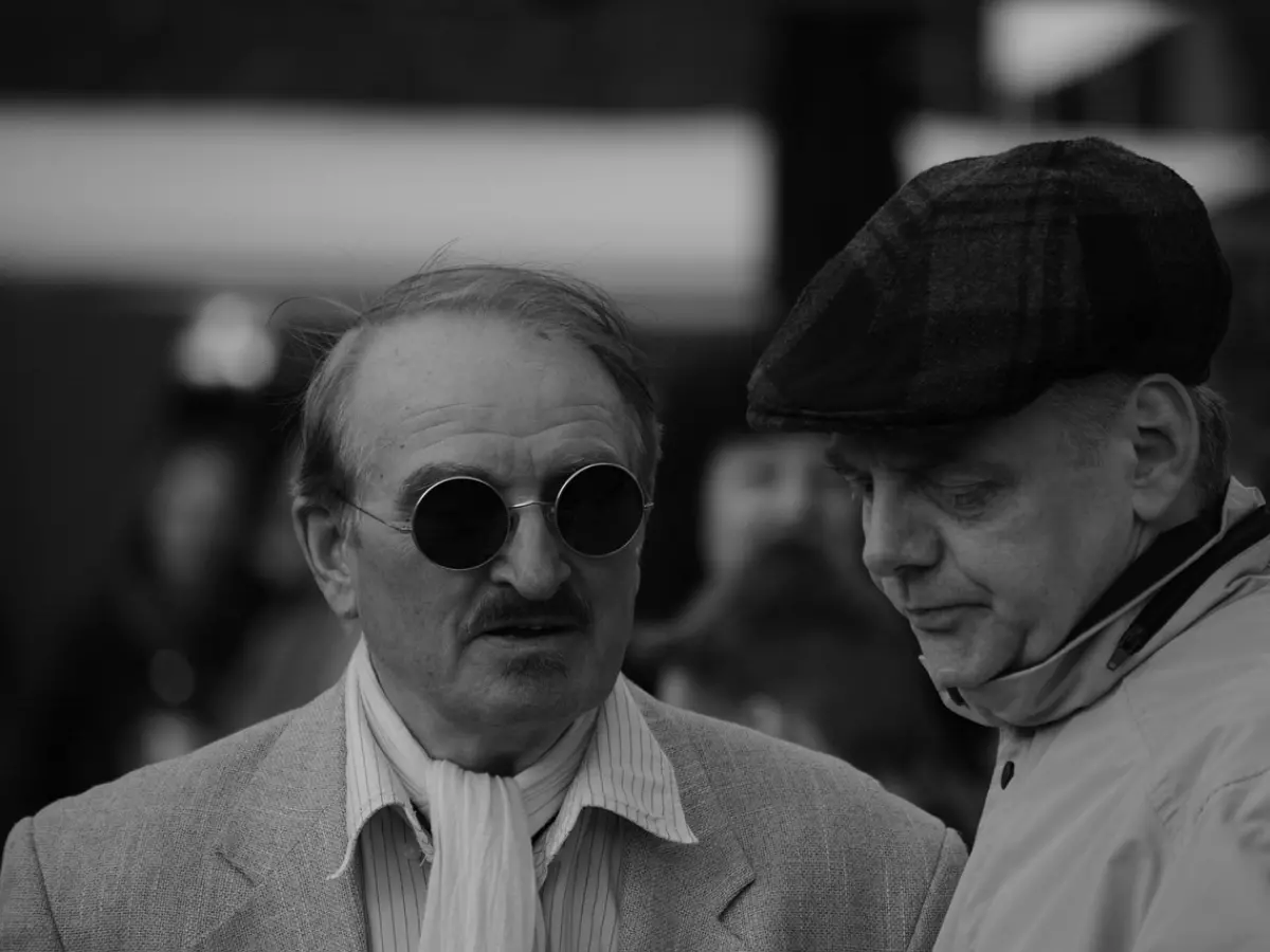 Two men are talking in a serious conversation. One man is wearing sunglasses and a scarf, and the other is wearing a cap. They are both looking at each other.