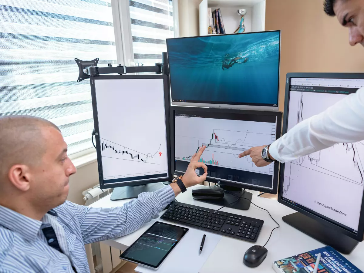 Two men in a office setting, looking at and pointing to screens displaying charts.