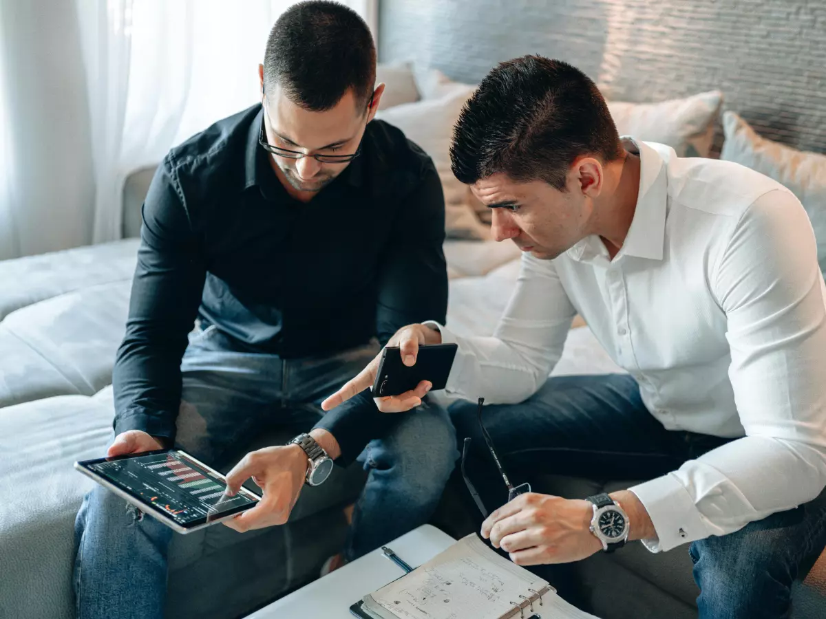 Two men sit on a couch, looking at a tablet and discussing something.