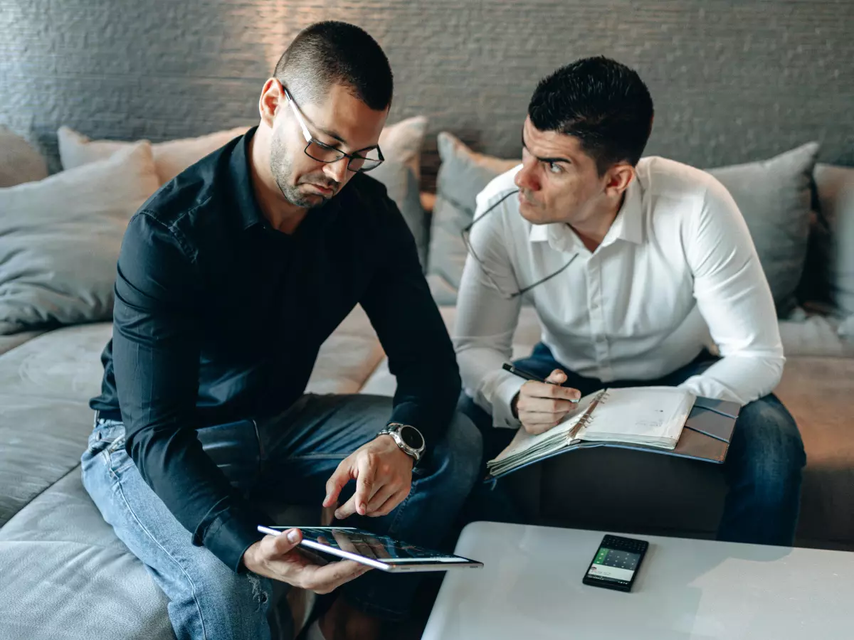 Two men are sitting on a couch, looking at a tablet. One is pointing at the screen while the other is writing in a notebook. There is a phone and a notepad on the coffee table.