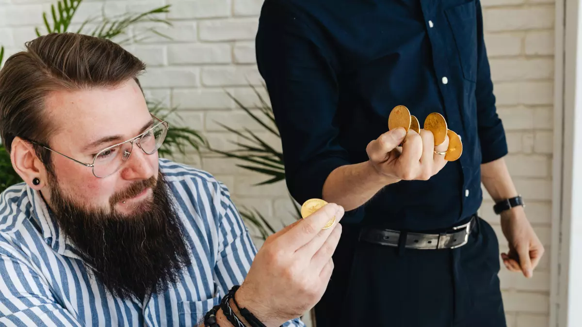 A man with a beard is holding a gold coin in his hand. He is sitting at a table. Another person is standing in the background, their hand is out of frame, holding what seems like another gold coin.