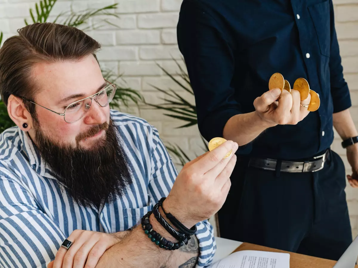 A man with a beard is holding a gold coin in his hand. He is sitting at a table. Another person is standing in the background, their hand is out of frame, holding what seems like another gold coin.