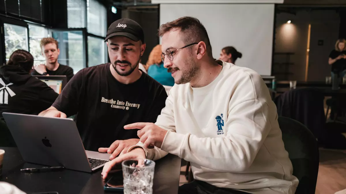 Two men are working on a laptop in a cafe, one pointing at the screen. 