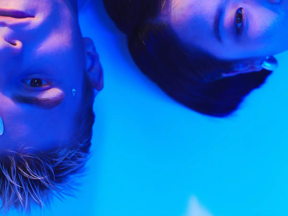 A man and a woman are lying on their backs in a blue-lit room, their eyes are open and staring directly at the camera. The man has blonde hair and the woman has dark hair. The image is taken from a low angle, so the viewer is looking up at the subjects. 