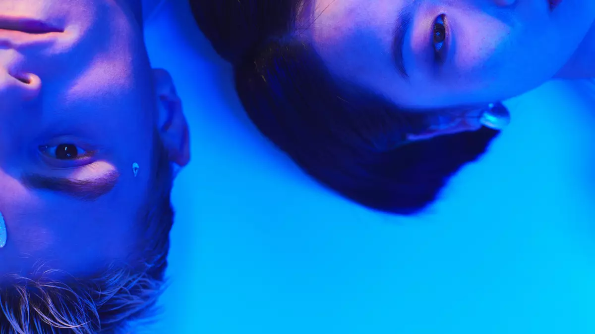 A man and a woman are lying on their backs in a blue-lit room, their eyes are open and staring directly at the camera. The man has blonde hair and the woman has dark hair. The image is taken from a low angle, so the viewer is looking up at the subjects. 