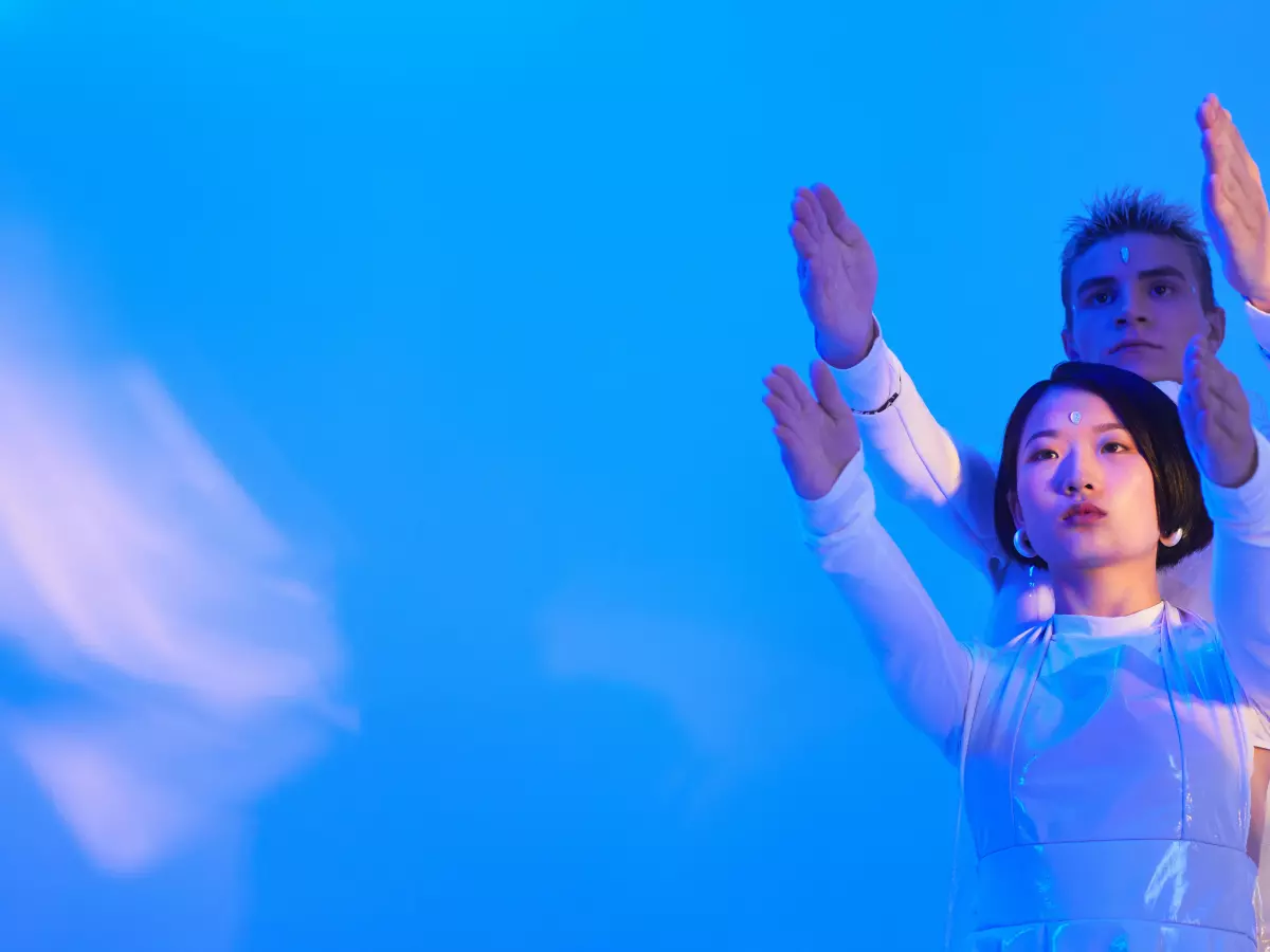 Two figures, one in white and the other in black, stand with their arms raised against a bright blue backdrop. The figure in white appears to be slightly translucent.