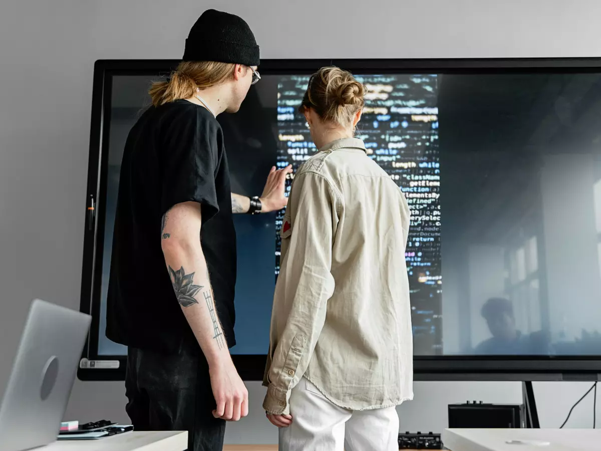 Two people are standing in front of a large screen, looking at code. 