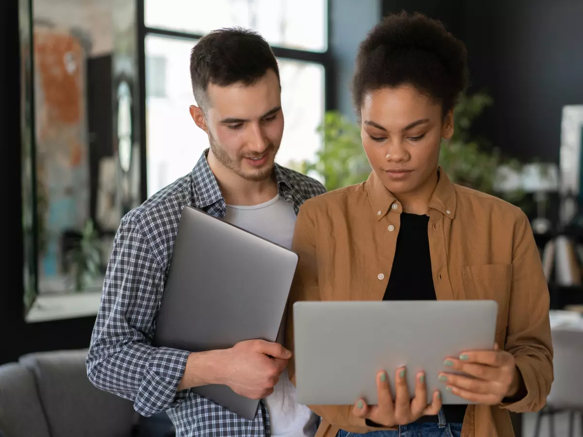 Two people, a man and a woman, are looking at a laptop. The man is standing and holding a closed laptop, while the woman is sitting and holding the open laptop. They are both looking at the screen.