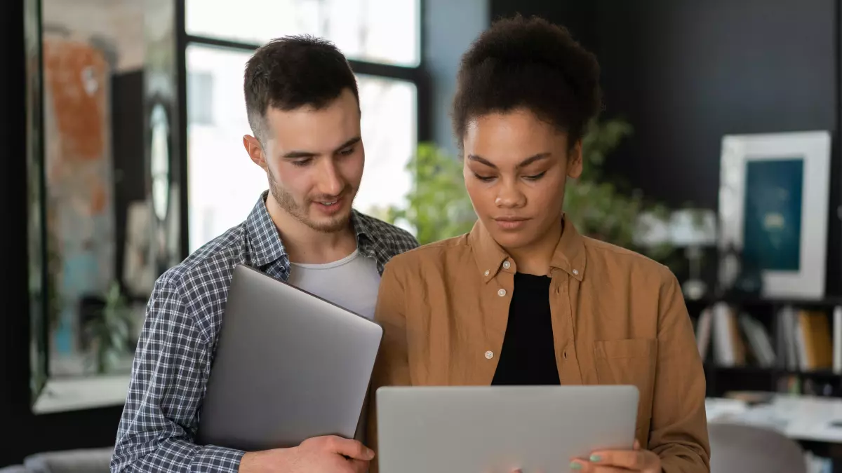 Two people, a man and a woman, are looking at a laptop. The man is standing and holding a closed laptop, while the woman is sitting and holding the open laptop. They are both looking at the screen.