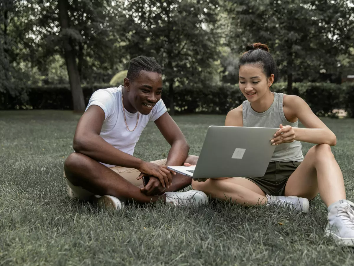 Two young people are sitting on the grass in a park and looking at a laptop.