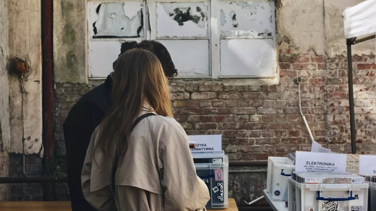 A young couple is browsing a selection of records at an outdoor market. The woman is looking at a crate of records, while the man is looking at a label. 