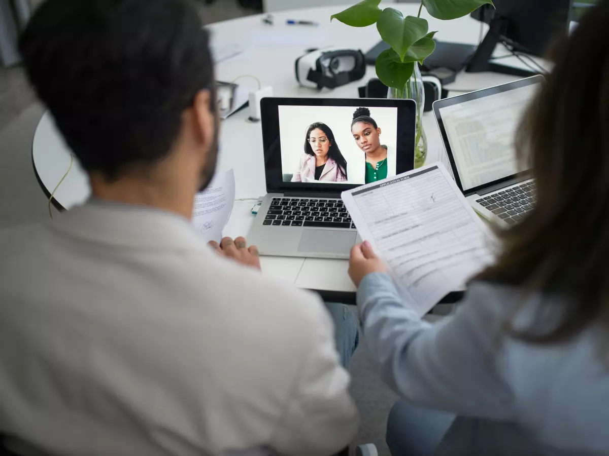 Two people look at a laptop screen during a video call interview, one is holding a resume.