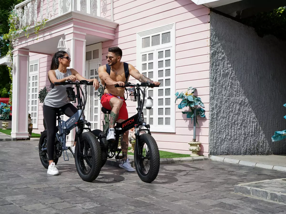 A couple riding electric bicycles on a path in front of a pink building.