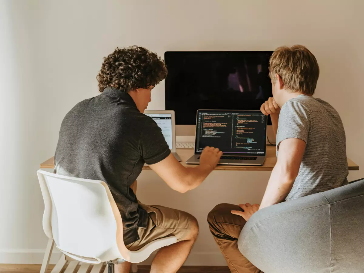 Two young men sit at a desk facing a computer screen, one pointing to the screen with his finger.