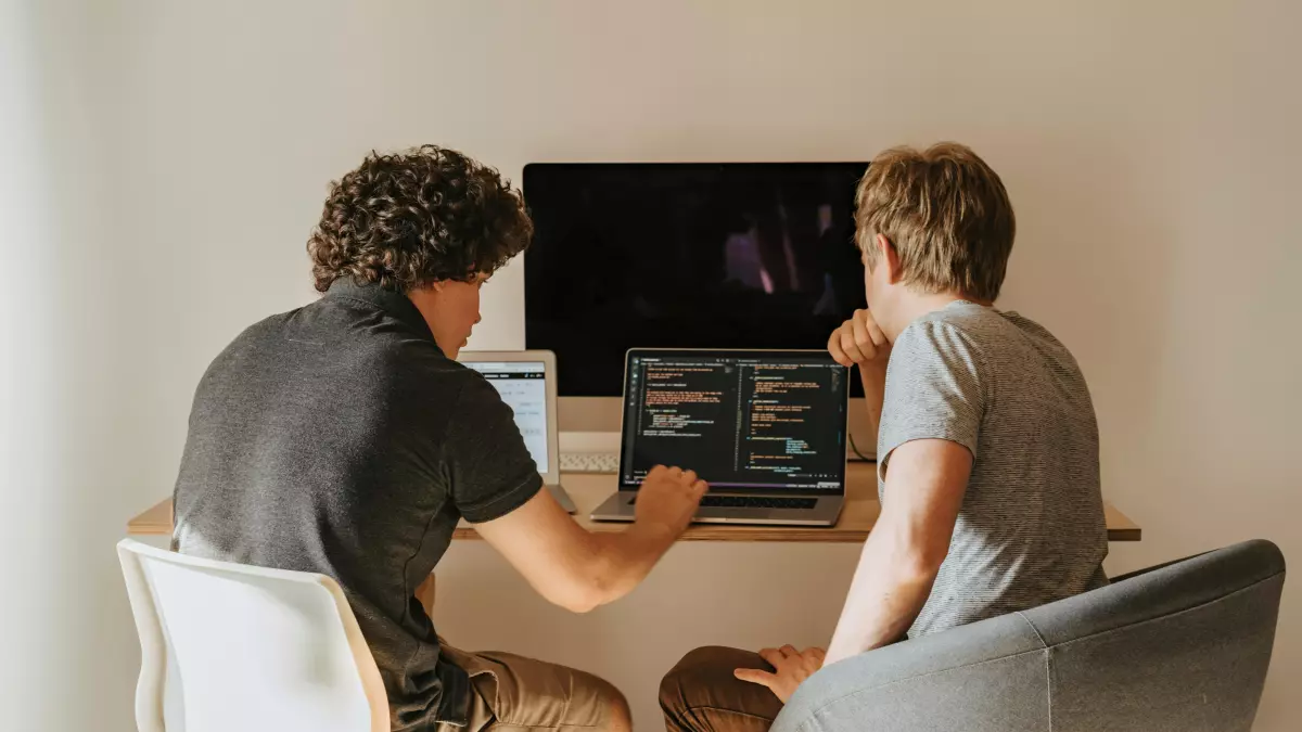 Two young men sit at a desk facing a computer screen, one pointing to the screen with his finger.