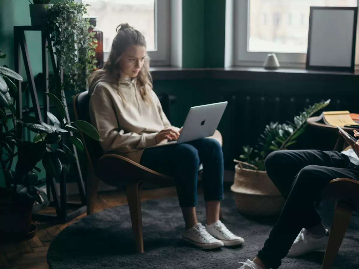 Two individuals are working on their laptops in a cozy home setting, with one person focused on their screen while the other looks at a document.