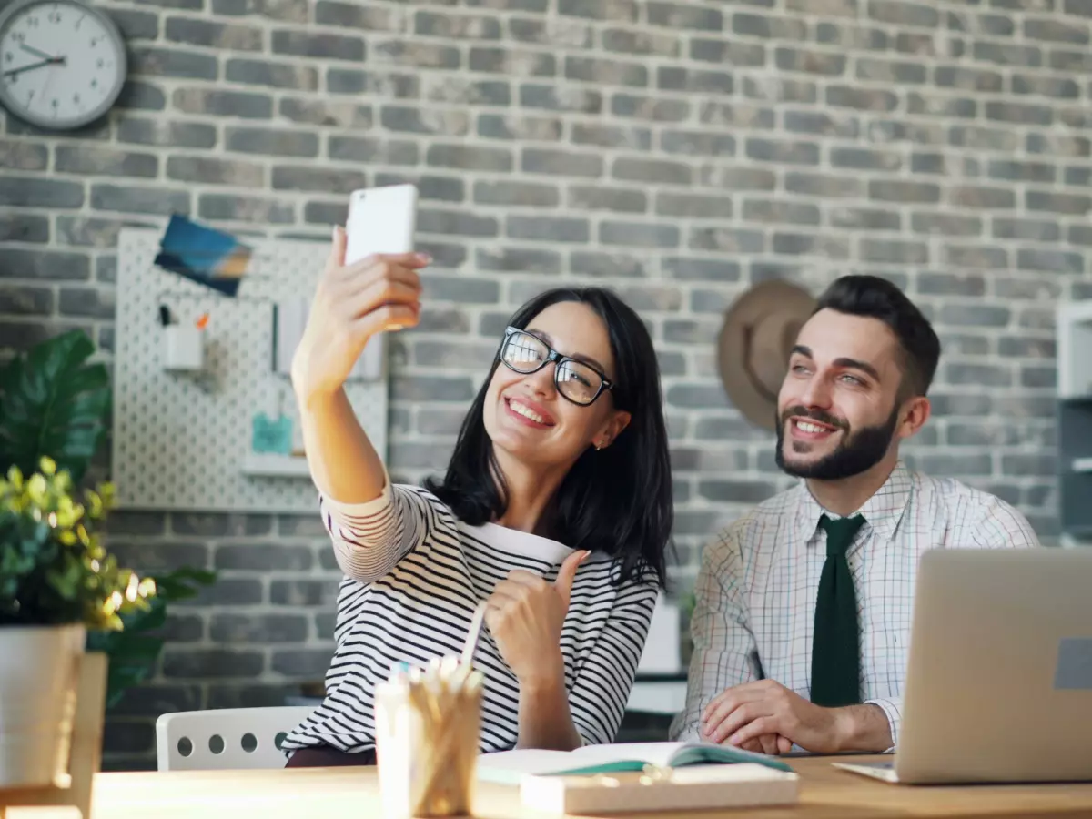 A man and a woman are taking a selfie with their iPhones. The man is using a laptop.