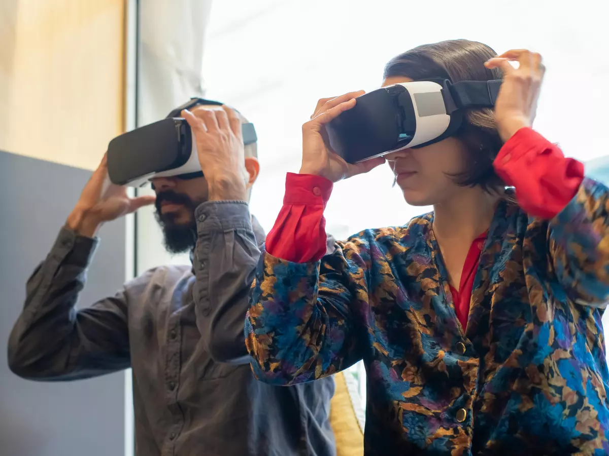 Two people wearing VR headsets are looking at a laptop screen. They are in a cafe or office setting.