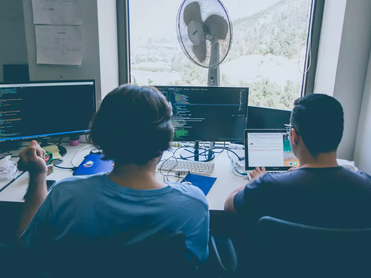 Two people are sitting at a desk in front of computers. The person in the background is wearing a blue shirt and the person in the foreground is wearing a black shirt. They are both looking at the computers and coding. There is a fan in the window.