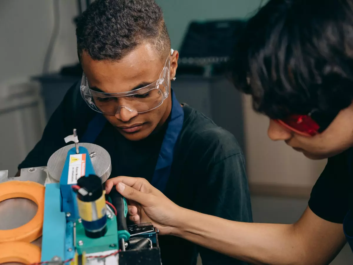 Two people are working on a robotic device, one holding a soldering iron and the other looking closely at the device.