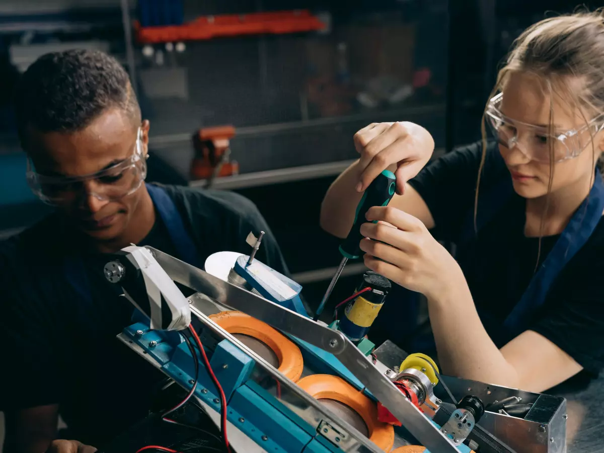 Two people, a man and a woman, are working on a robotic device. They are wearing safety glasses and focused on their task. The setting appears to be a workshop.