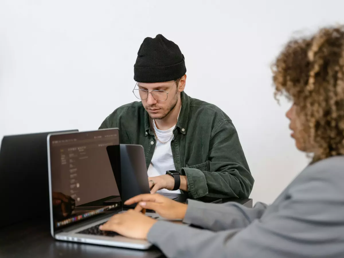 A man in a green jacket and a black beanie sits in front of a laptop, typing, while a woman looks over his shoulder. Both appear focused, with a serious expression.