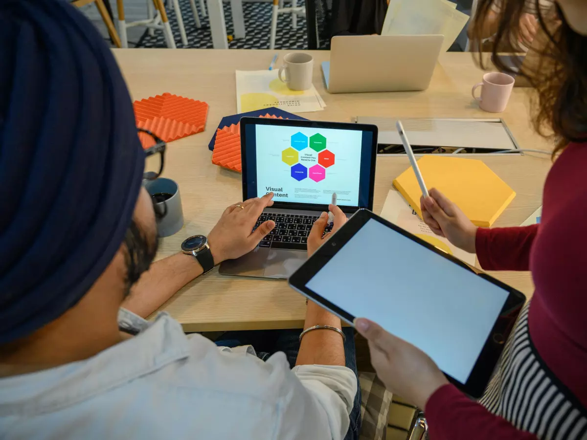 Two people working together at a desk. One person is using a laptop and the other is using a tablet.