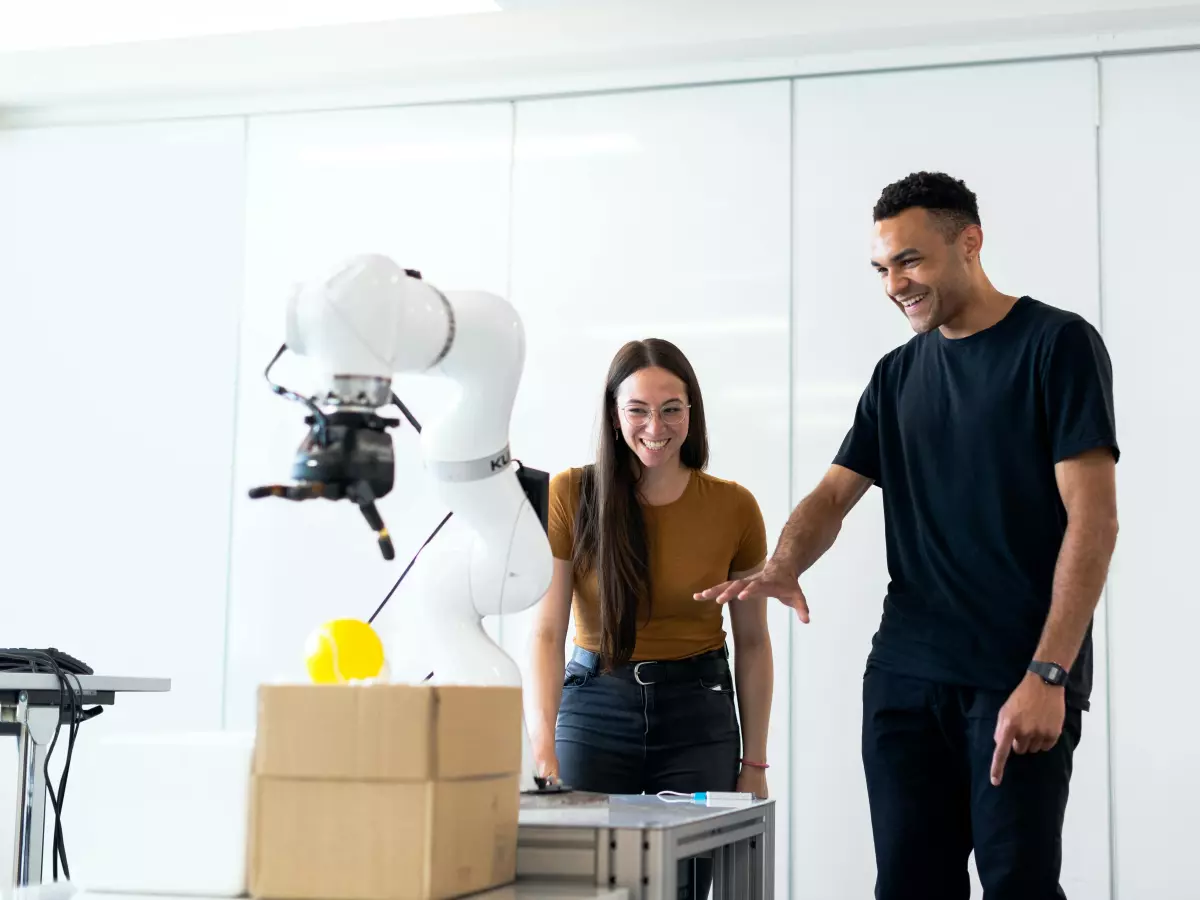 A man and a woman are standing in front of a robotic arm, the man is pointing at the robotic arm and the woman is looking at the robotic arm.