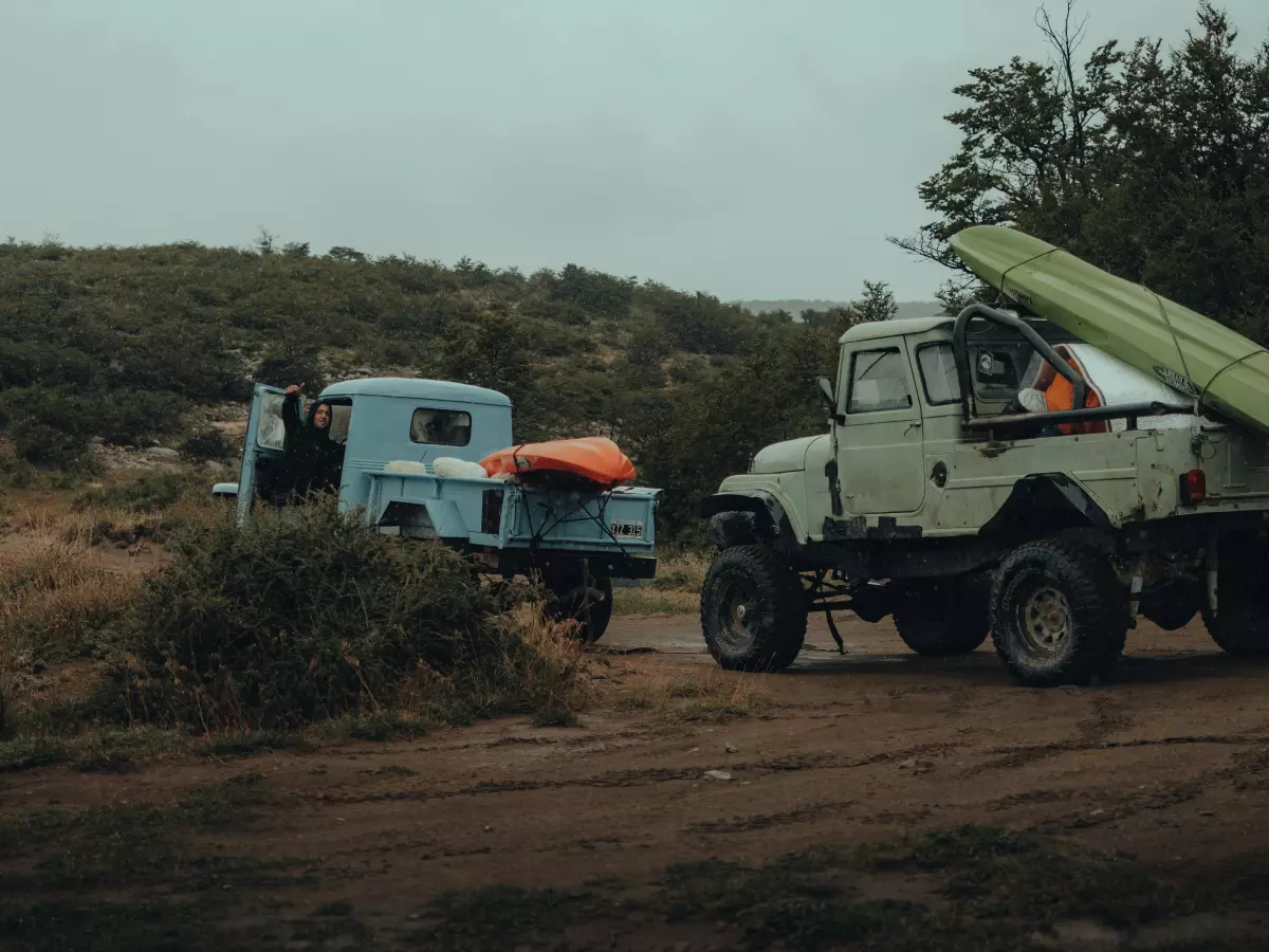 An off-road truck is parked on a dirt path, with a kayak strapped on the back. There is a person in the background.