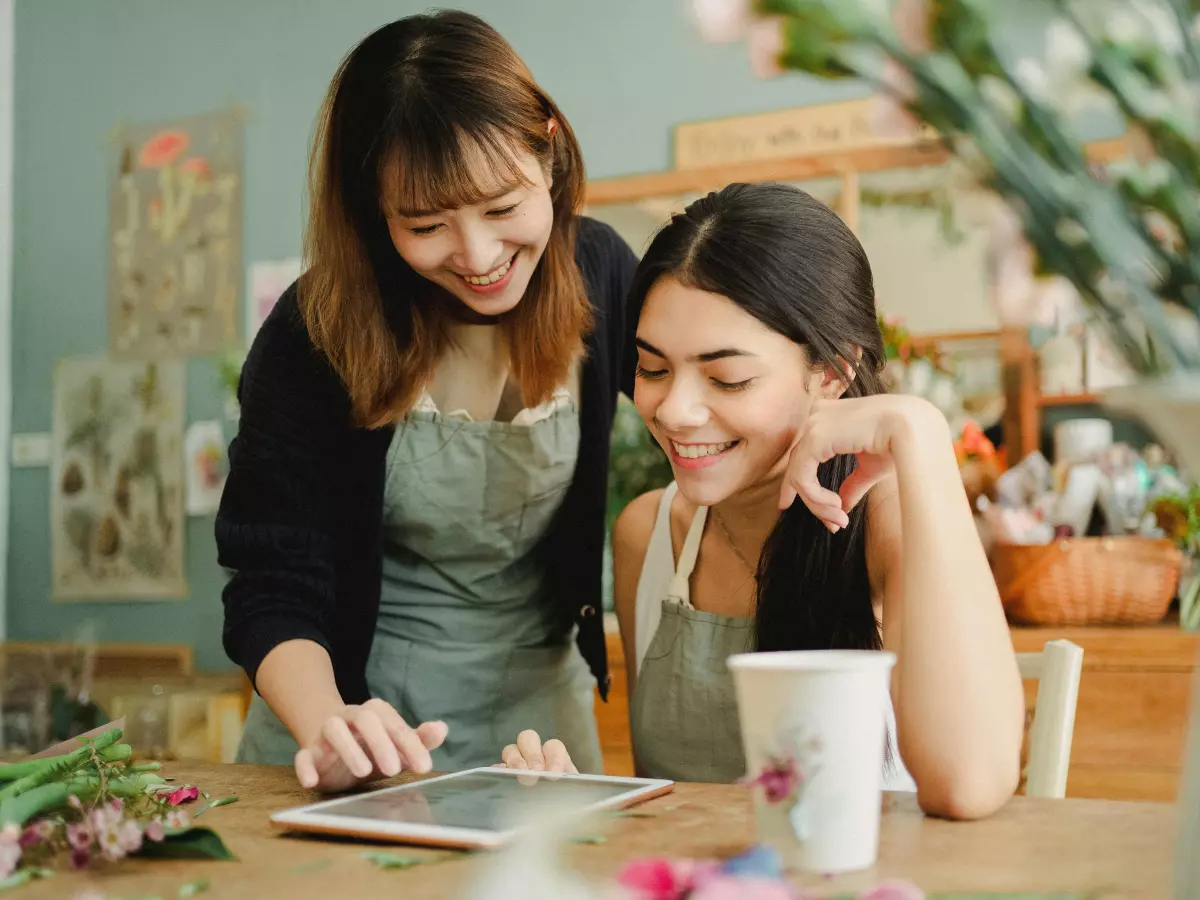 Two women are using an iPad in a flower shop. One is standing while the other is sitting and they are both smiling. 