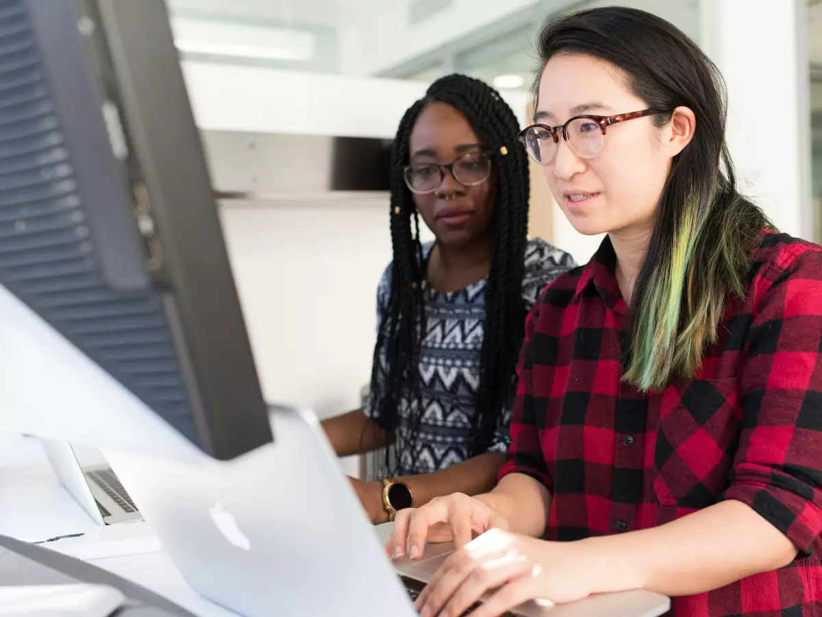 Two women, one black and one asian, are sitting at a desk and working on a computer. They are both looking at the screen and are focused on their work. The asian woman has on a red plaid shirt and black framed glasses. The black woman has on a patterned gray and black shirt. The photo is taken from a low angle, and the women are in the foreground, while the office is in the background.