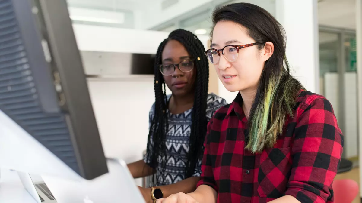 Two women, one black and one asian, are sitting at a desk and working on a computer. They are both looking at the screen and are focused on their work. The asian woman has on a red plaid shirt and black framed glasses. The black woman has on a patterned gray and black shirt. The photo is taken from a low angle, and the women are in the foreground, while the office is in the background.