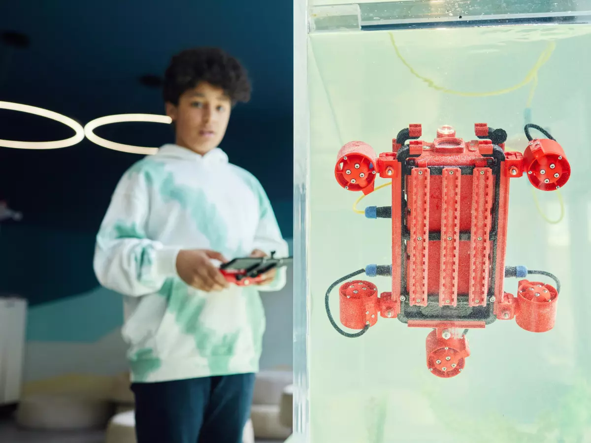 A young person, likely a teenager, operates a small, red robot contained in a glass tank. The person is holding a controller, and the robot is moving.