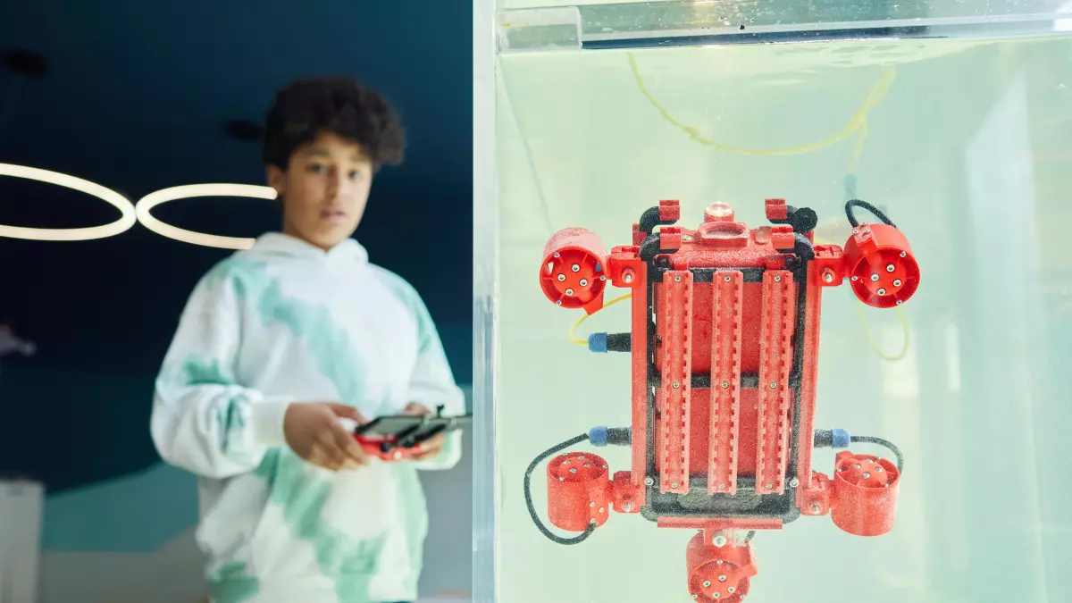 A young person, likely a teenager, operates a small, red robot contained in a glass tank. The person is holding a controller, and the robot is moving.