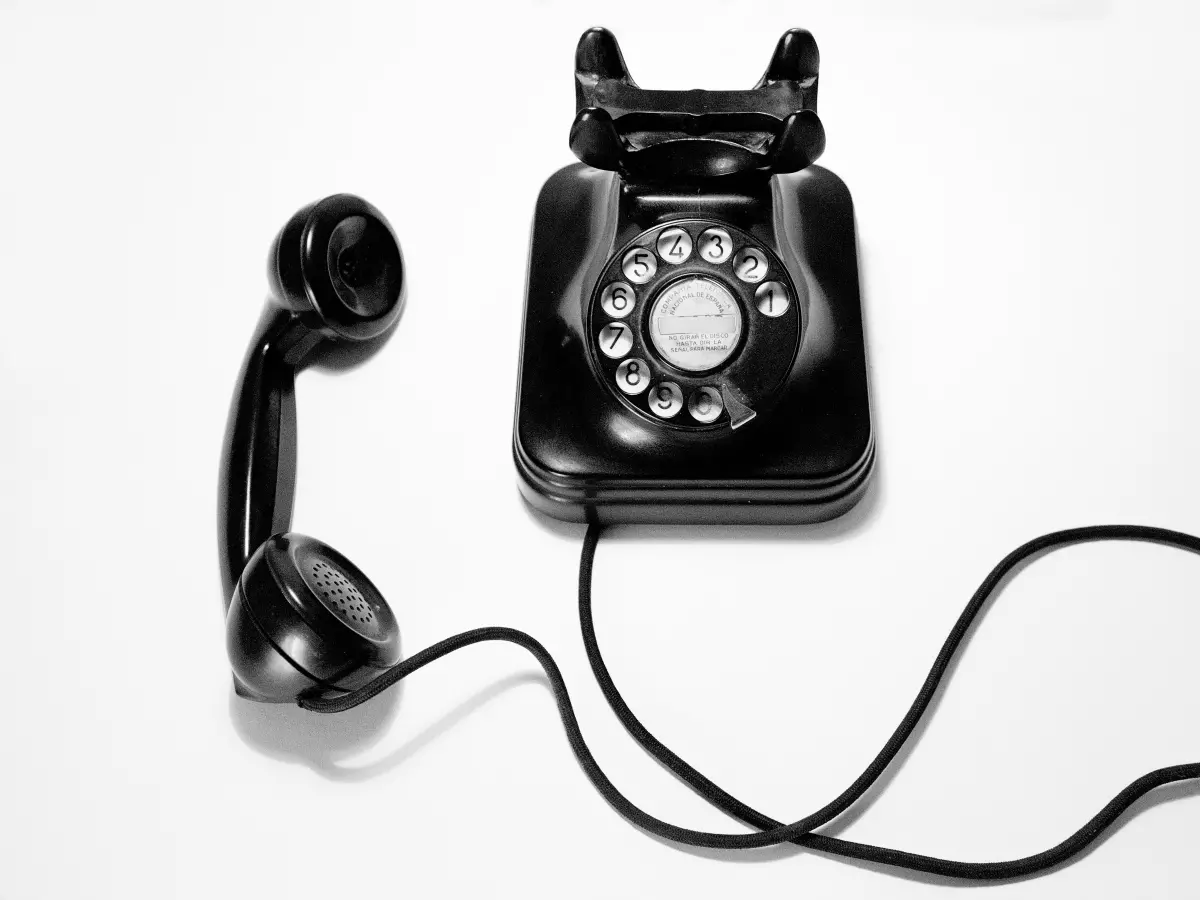 A black rotary phone with a white dial and a black handset lying on a white surface. The handset is connected to the phone by a black cord.