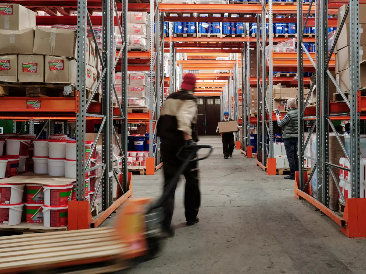 A person on a pallet truck in a warehouse aisle with shelves stacked high on both sides.