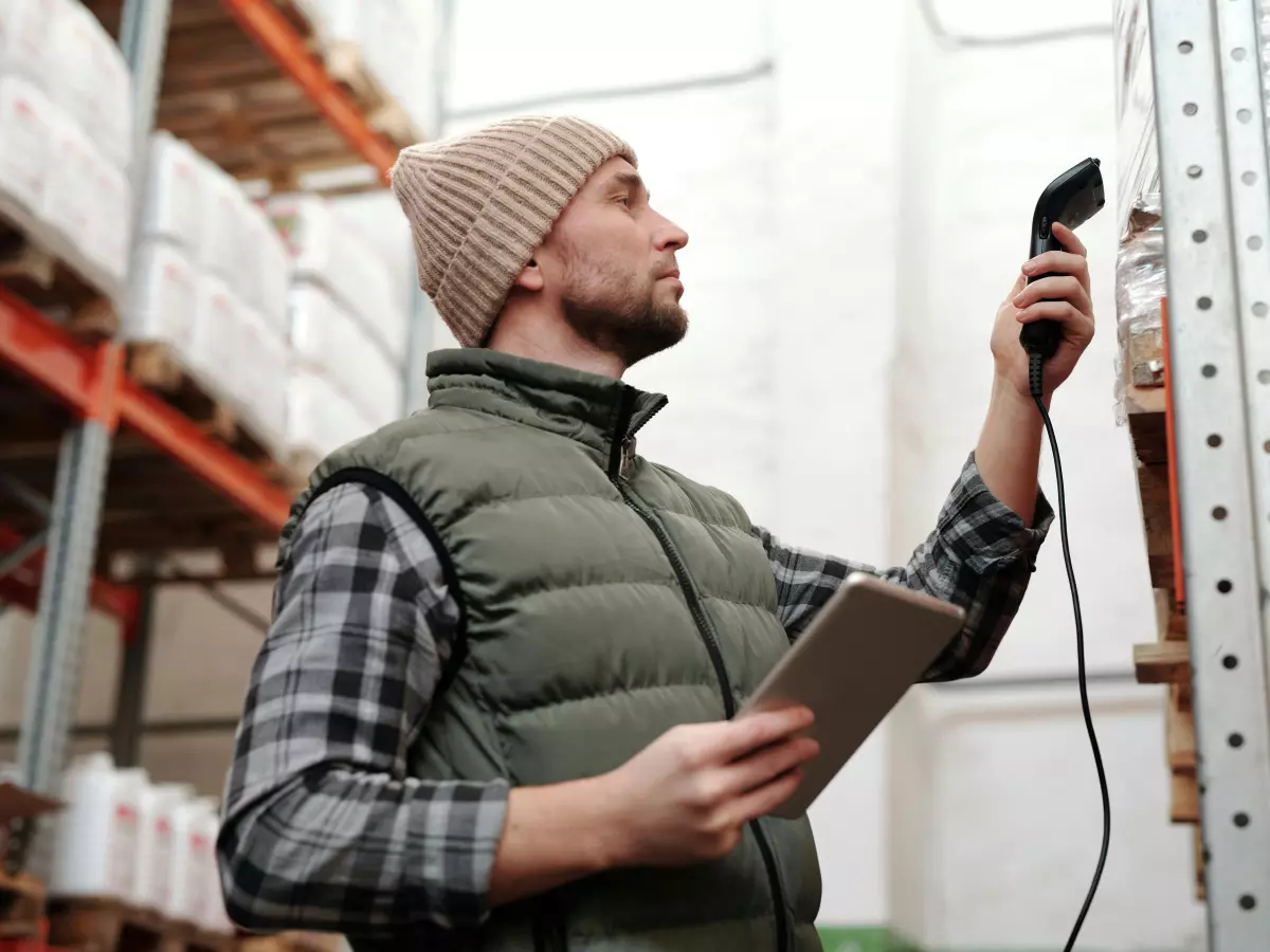 A man in a warehouse is scanning a barcode with a scanner and holding a tablet in the other hand. 