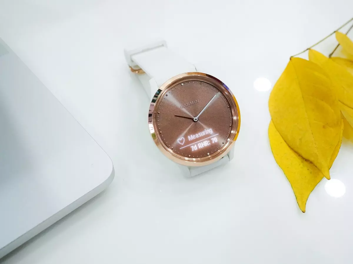 A white smartwatch with a gold face on a white surface, next to a laptop and yellow leaves.