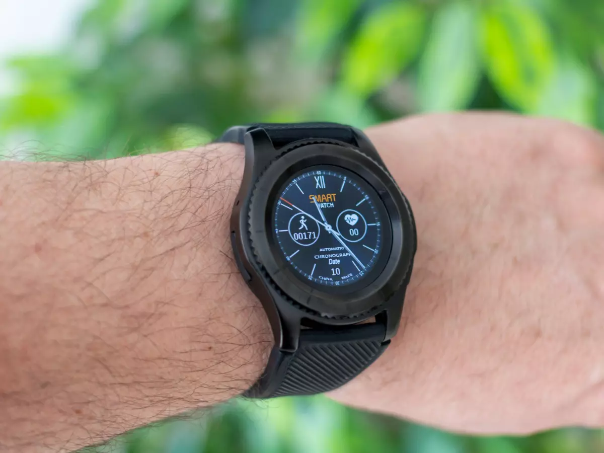 A person wearing a black smartwatch on their left wrist. The watch face is a digital one and shows the time, date, steps taken, heart rate, and battery life. 